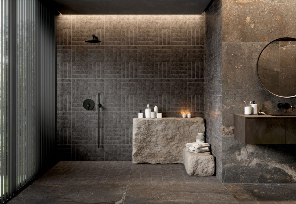 Modern bathroom with dark stone tiles, a sleek black showerhead, and a circular mirror. Minimalist design with candles and toiletries on stone surfaces.