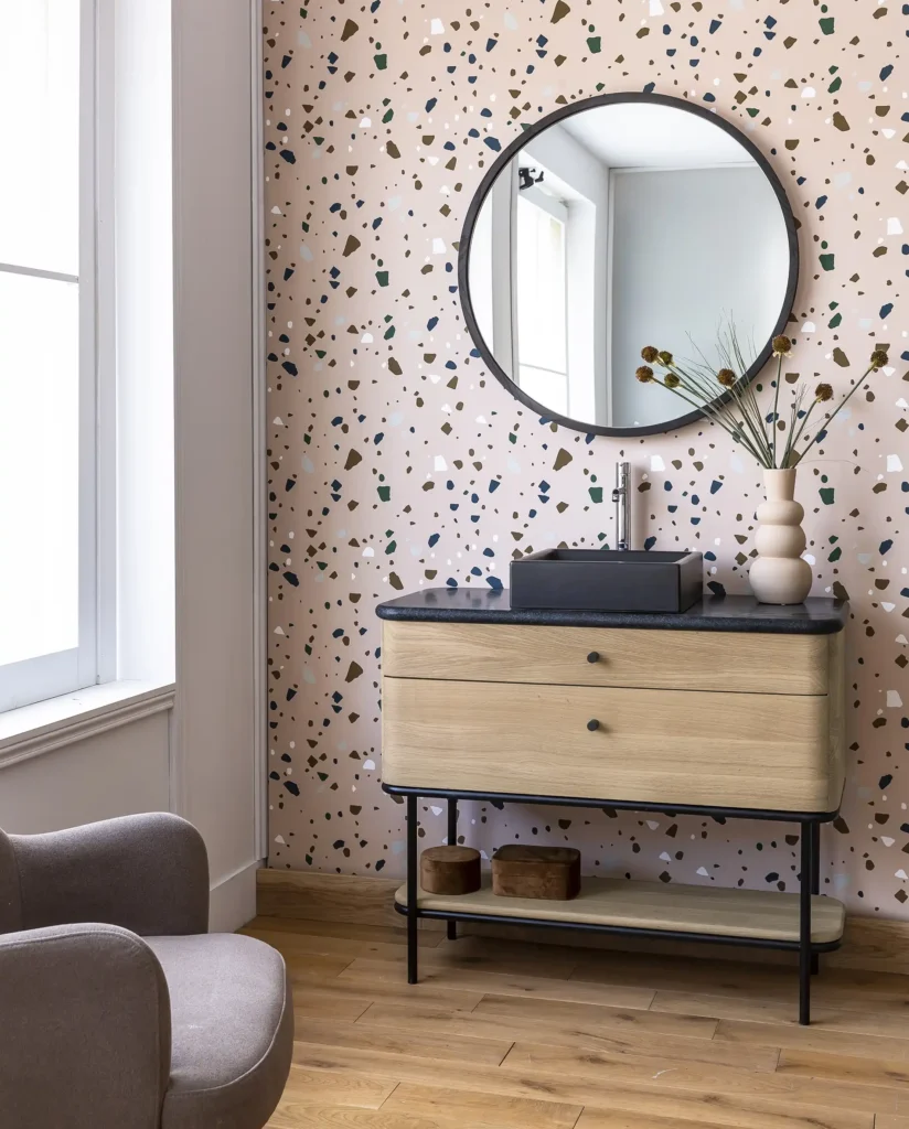 A modern bathroom features terrazzo wallpaper, a round mirror, and a wooden vanity with a black rectangular sink. A minimalist vase and chair add elegance.