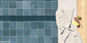 Top-down view of a pool edge with Colonial Blue square tiles, featuring a white towel, a refreshing summer drink with lemon, and wooden decking.