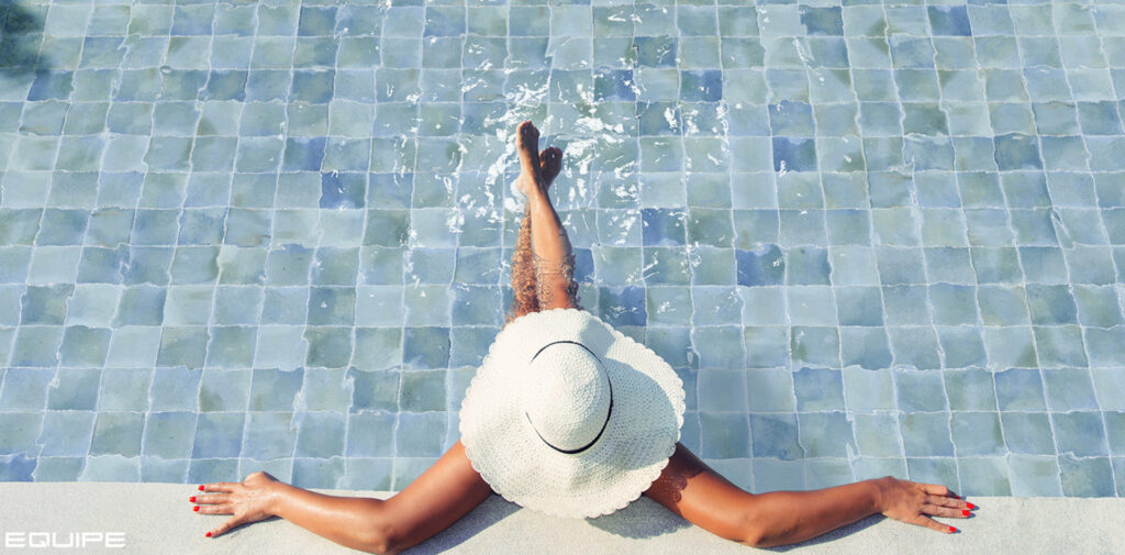 A person in a white sun hat relaxes at the edge of a pristine blue-tiled pool. Their legs are in the water, and their arms are stretched out, conveying tranquility.