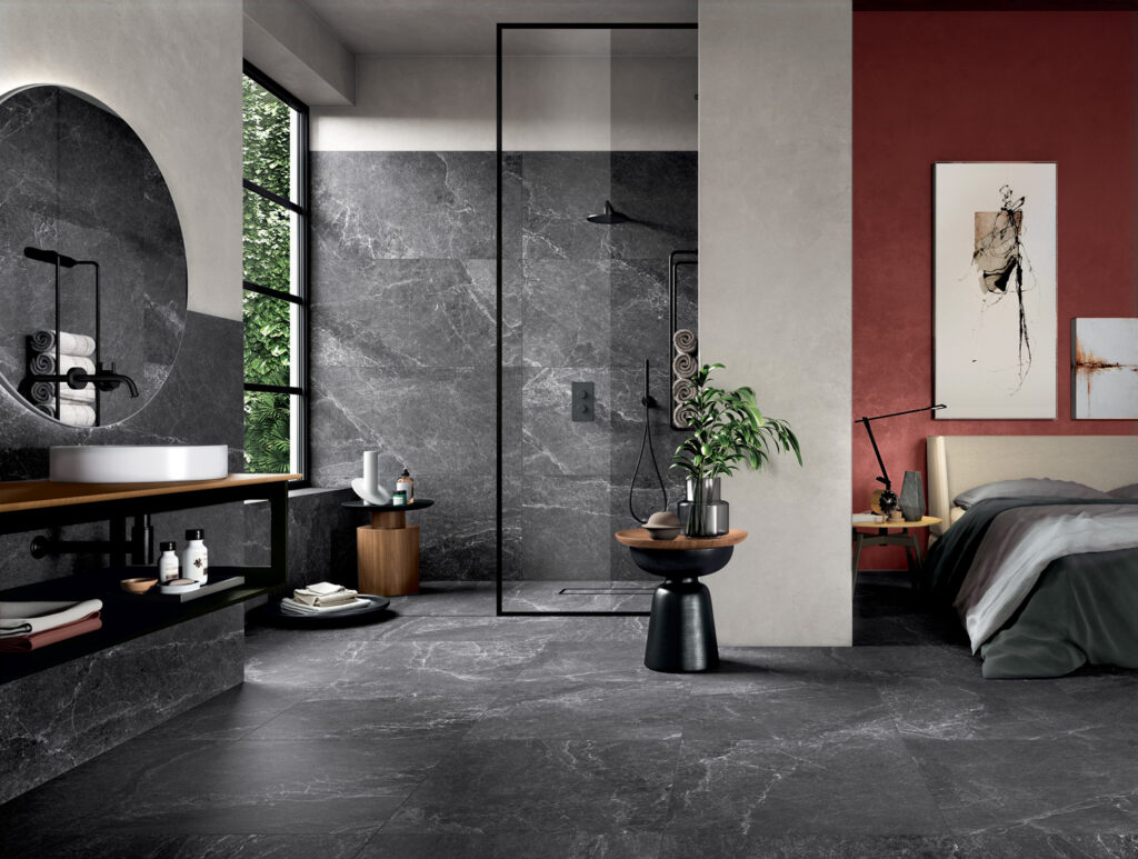 Modern bathroom and bedroom combo with a sleek black marble shower, large round mirror, dark gray flooring, red accent wall, and greenery for a fresh look.