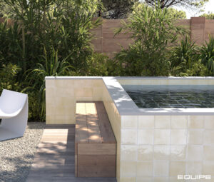 Outdoor view of a modern raised spa pool with cream-colored, textured Zellige-style tiles by Equipe, featuring a wooden bench and white accent chair in a lushly landscaped backyard.